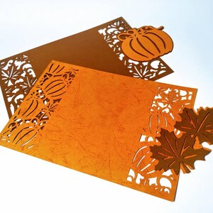 May include: Two orange and brown paper placemats with a cut-out design of pumpkins, leaves, and flowers. The placemats are on a white background.