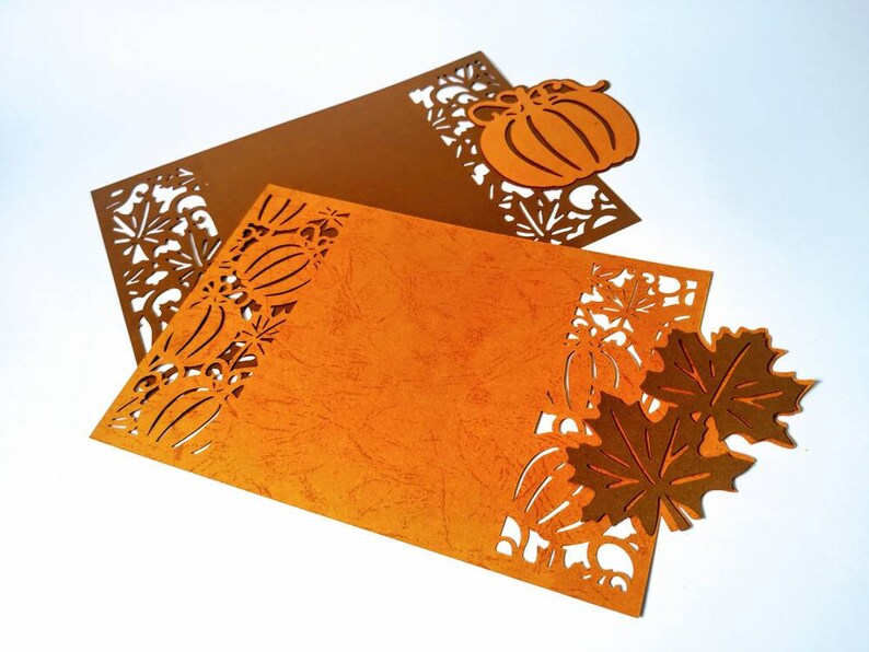 May include: Two orange and brown paper placemats with a cut-out design of pumpkins, leaves, and flowers. The placemats are on a white background.