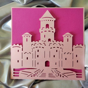 May include: A pink paper card with a white castle cut out. The castle has a tall tower in the center and two smaller towers on either side. The castle has a bridge leading to the front door.