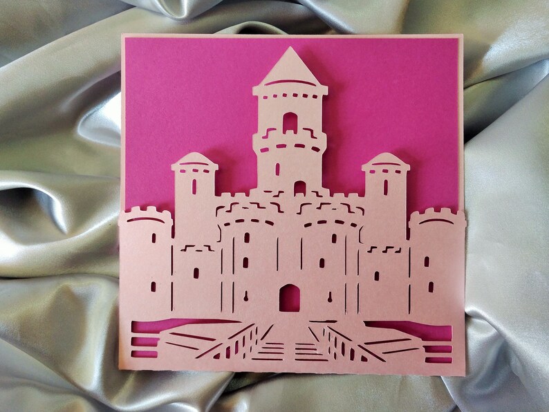 May include: A pink paper card with a white castle cut out. The castle has a tall tower in the center and two smaller towers on either side. The castle has a bridge leading to the front door.