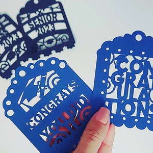 May include: Three blue paper cut-out tags with white lettering. One tag says "Class of 2023", another says "Senior 2023", and the third says "Congratulations You Did It".