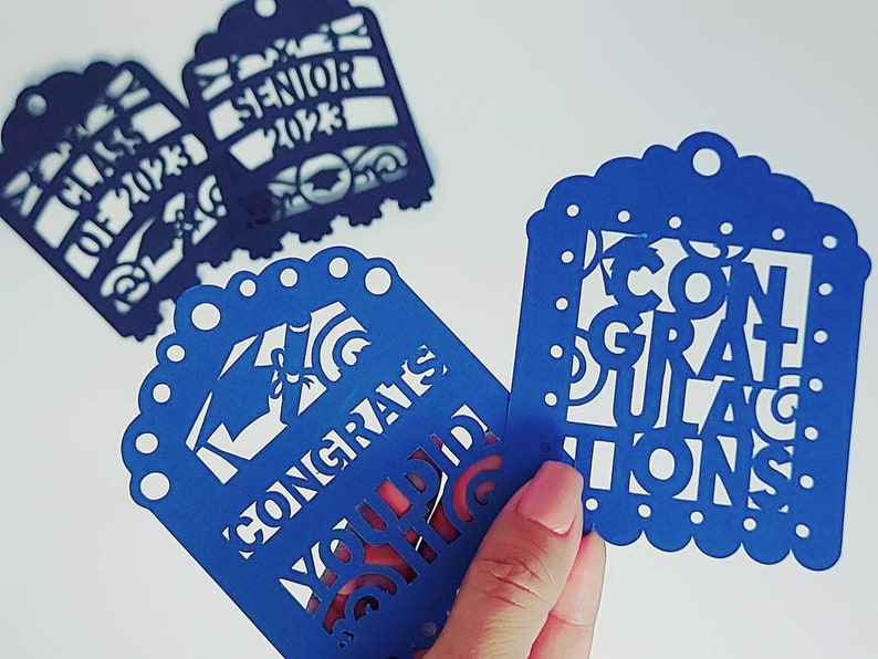 May include: Three blue paper cut-out tags with white lettering. One tag says "Class of 2023", another says "Senior 2023", and the third says "Congratulations You Did It".