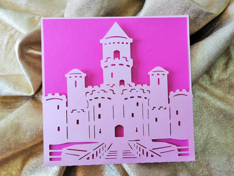May include: A white paper cut-out of a castle with a pink background. The castle has three towers and a drawbridge.