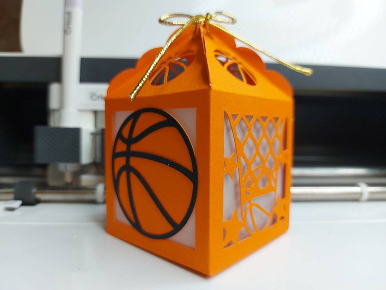 May include: Orange paper favor box with a basketball design and a gold ribbon. The box has a cut-out design of a basketball hoop and net.