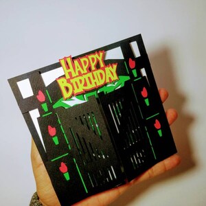 May include: A black and green 3D pop-up card with a gate design. The card has a red and yellow "Happy Birthday" message on the front.