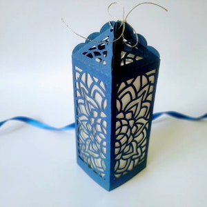 May include: A blue paper gift box with a delicate, intricate cut-out design. The box is open and has a white interior. A silver ribbon is tied around the top of the box.