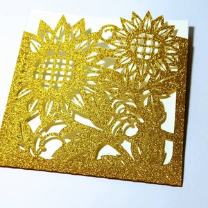 May include: Gold glitter card with a cut-out design of two sunflowers. The card is folded in half and has a white interior.