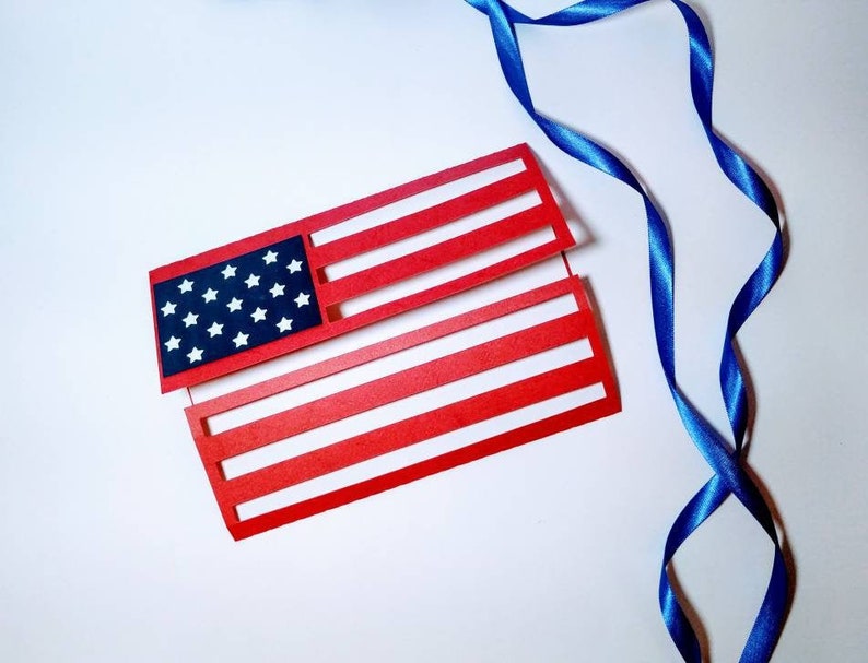 May include: A red, white, and blue paper American flag with a blue ribbon on a white background.