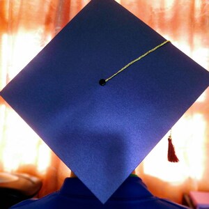 Puede incluir: Un gorro de graduaci&oacute;n azul con una borla dorada y un bot&oacute;n negro en la parte superior. El gorro lo lleva puesto una persona con una camisa azul.