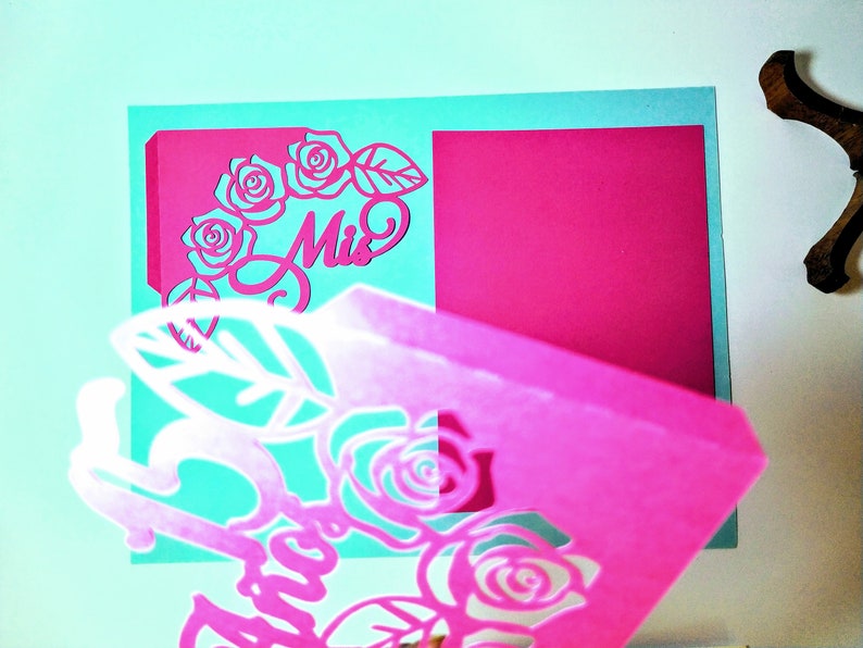 May include: A pink and teal paper craft with a cut-out design of roses and the word "Miss".