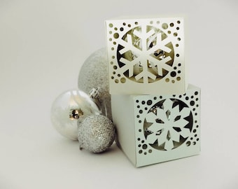 May include: Two white cardboard boxes with snowflake cutouts. The boxes are stacked on top of each other. There are three silver Christmas ornaments in front of the boxes.