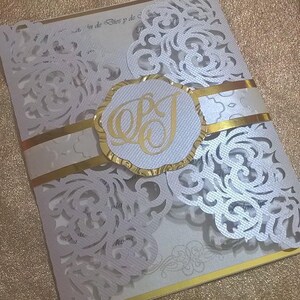 May include: White wedding invitation with a gold trim and a white lace overlay. The invitation has a gold circle with the initials "EJ" in the center.