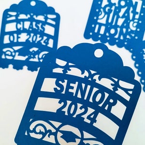 May include: Four blue paper cut-out tags with graduation themes. One tag says "Class of 2024", another says "Senior 2024", another says "Congratulations", and the last one says "You Did It!"