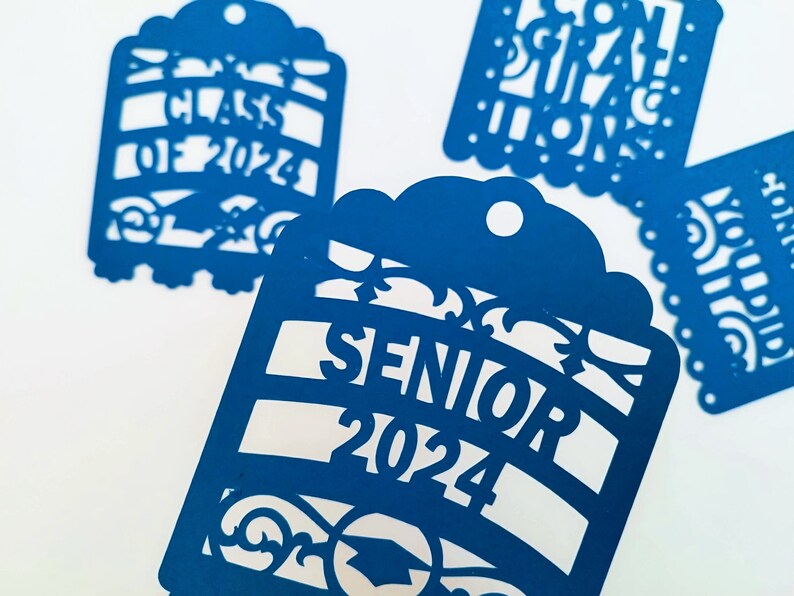 May include: Four blue paper cut-out tags with graduation themes. One tag says "Class of 2024", another says "Senior 2024", another says "Congratulations", and the last one says "You Did It!"