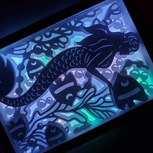 May include: A black silhouette of a mermaid with a long flowing tail, surrounded by a white and blue patterned design. The design includes fish and seaweed. The image is illuminated from behind, creating a glowing effect.