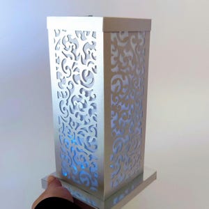 Op de afbeelding: Een zilveren rechthoekige lamp met ingewikkelde, wervelende uitgesneden patronen. De lamp straalt een zacht blauw licht uit, wat een decoratief en sfeervol lichteffect creëert. De lamp staat op een vierkante basis, ongeveer 30 cm hoog.