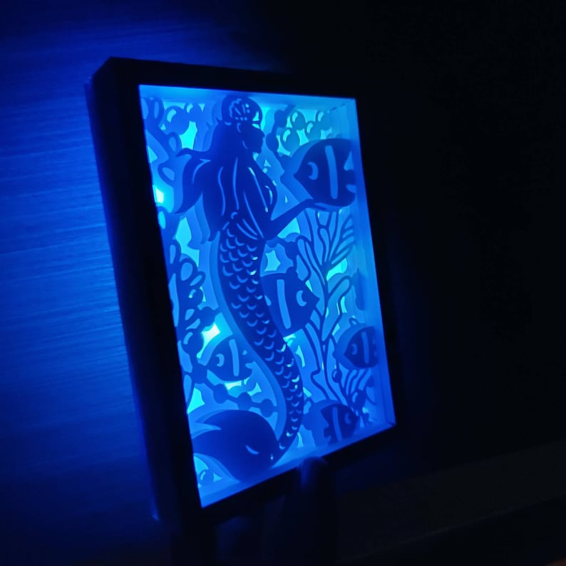 May include: A white night light with a blue glow, featuring a cutout design of a mermaid with flowing hair and a tail, surrounded by fish and coral.