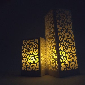 May include: Three white decorative candle holders with intricate cut-out designs. The holders are illuminated from within, casting a warm glow.