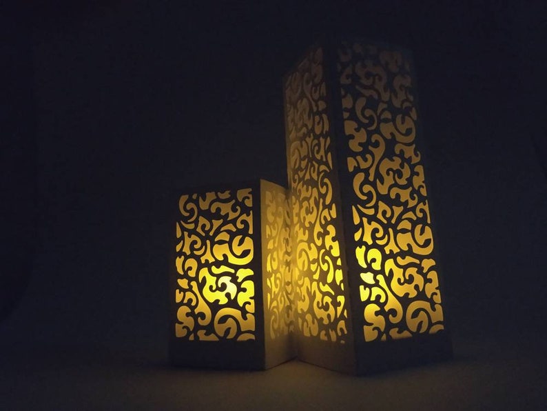 May include: Three white decorative candle holders with intricate cut-out designs. The holders are illuminated from within, casting a warm glow.