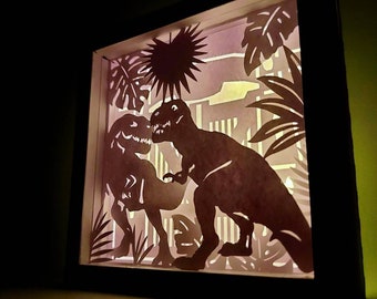 Pode incluir: Uma caixa de luz de papel recortado e emoldurada, com uma cena pré-histórica. Duas silhuetas de dinossauros enfrentam-se, rodeadas por folhagens estilizadas e um sol. A obra de arte tem um fundo gradiente.