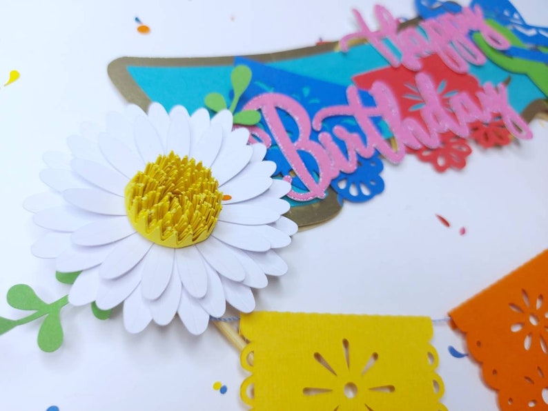 May include: A paper banner with the words "Happy Birthday" in pink letters. The banner is decorated with colorful paper flowers, leaves, and confetti.