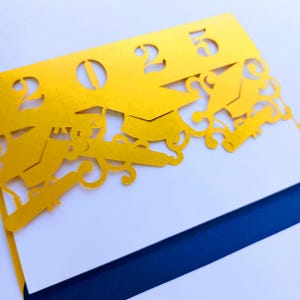 以下が含まれることがあります： 2023年が切り抜かれた金色のカードで、卒業帽と卒業証書が装飾的なボーダーになっています。カードは半分に折り畳まれており、ネイビーブルーの裏地が付いています。