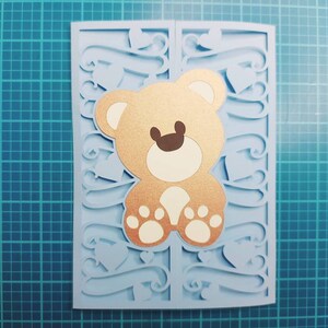 May include: A light blue card with a cut-out design featuring hearts and swirls. A brown teddy bear with white paws is in the center of the card.