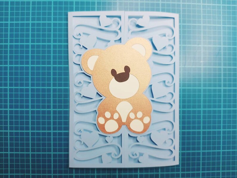May include: A light blue card with a cut-out design featuring hearts and swirls. A brown teddy bear with white paws is in the center of the card.