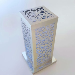 May include: A silver rectangular lamp with decorative cut-out patterns. The lamp has a square base and top, with a light blue glow visible through the cut-outs. The design is suitable for home decor, with a height of approximately 30 cm.