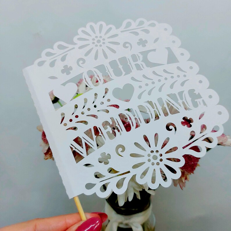 May include: A white paper cake topper with the words "OUR WEDDING" cut out in a decorative font. The topper has a scalloped edge with floral and heart cutouts. A wooden skewer is attached to the bottom.