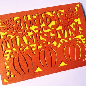 Puede incluir: Tarjeta naranja con fondo amarillo y un dise&ntilde;o recortado de calabazas y las palabras "Feliz D&iacute;a de Acci&oacute;n de Gracias".