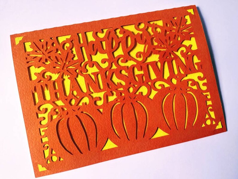 Puede incluir: Tarjeta naranja con fondo amarillo y un dise&ntilde;o recortado de calabazas y las palabras "Feliz D&iacute;a de Acci&oacute;n de Gracias".