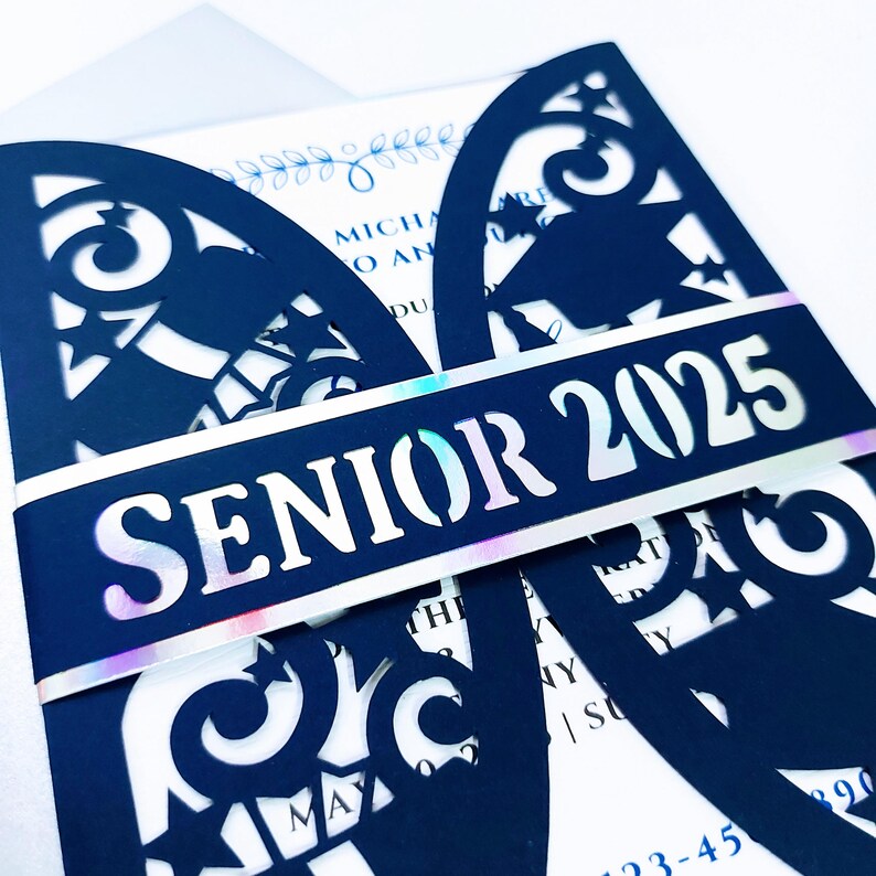May include: A navy blue graduation invitation with a laser cut design featuring stars and a graduation cap. The invitation has a white band with iridescent foil and the text "Senior 2025".