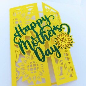 Op de afbeelding: Een gele kaart met een groene glitter-uitsparing van de woorden "Happy Mother's Day". De kaart is versierd met gele papieren uitsnijdingen van zonnebloemen.