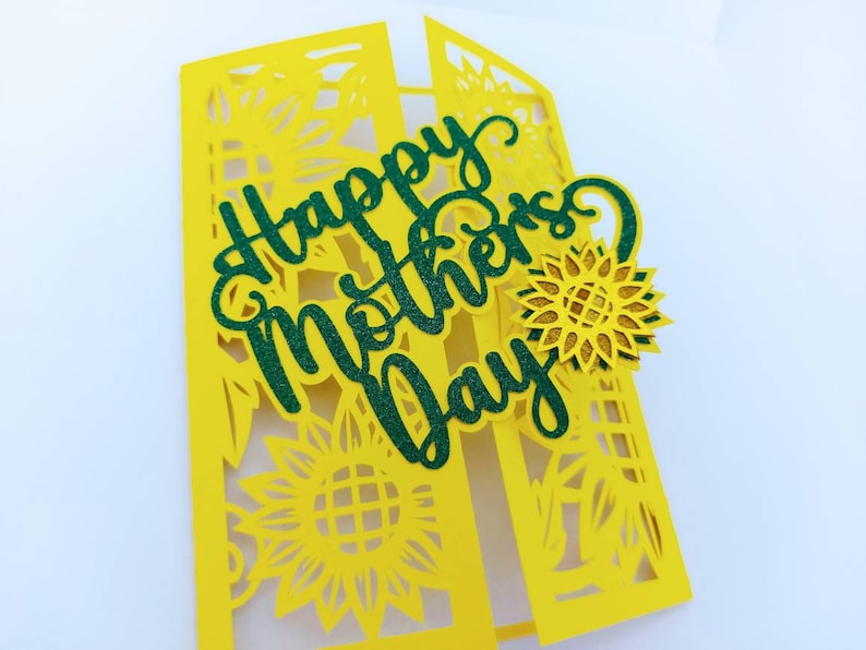 Op de afbeelding: Een gele kaart met een groene glitter-uitsparing van de woorden "Happy Mother's Day". De kaart is versierd met gele papieren uitsnijdingen van zonnebloemen.