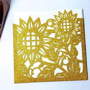 May include: Gold glitter laser cut card with a sunflower design. The card is folded in half and has a cutout design of sunflowers and leaves.