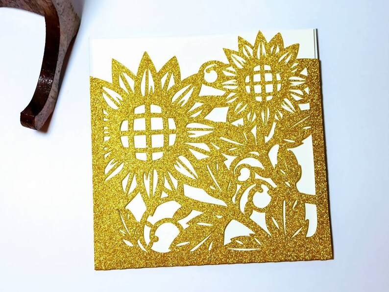 May include: Gold glitter laser cut card with a sunflower design. The card is folded in half and has a cutout design of sunflowers and leaves.