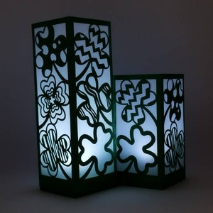 Puede incluir: Dos faroles de papel con diseños en blanco y negro y un borde verde azulado. Los faroles están iluminados desde el interior, proyectando un suave resplandor.