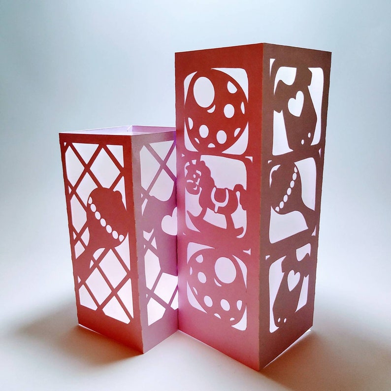May include: Pink paper lantern with cut-out designs of a rocking horse, a ball, a rattle, and a onesie. The lantern is made of multiple panels that are joined together.
