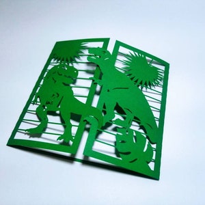 May include: Green paper card with a cut-out design of two dinosaurs. The dinosaurs are facing each other and are surrounded by a sunburst design.