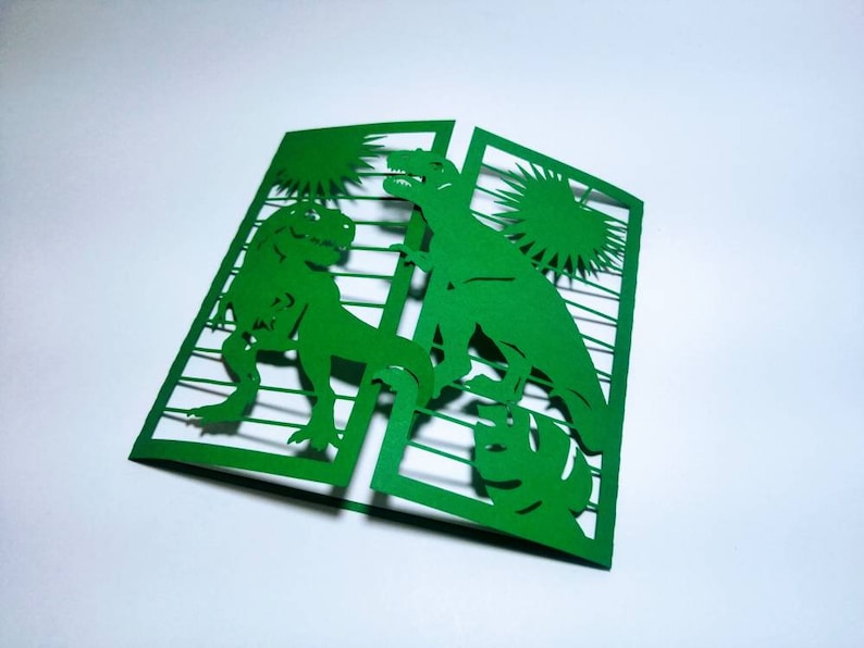 May include: Green paper card with a cut-out design of two dinosaurs. The dinosaurs are facing each other and are surrounded by a sunburst design.