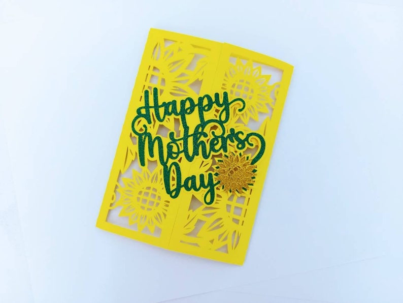 Op de afbeelding: Een gele kaart met een groene glitter-uitsparing van de woorden "Happy Mother's Day". De kaart is versierd met een zonnebloemontwerp.