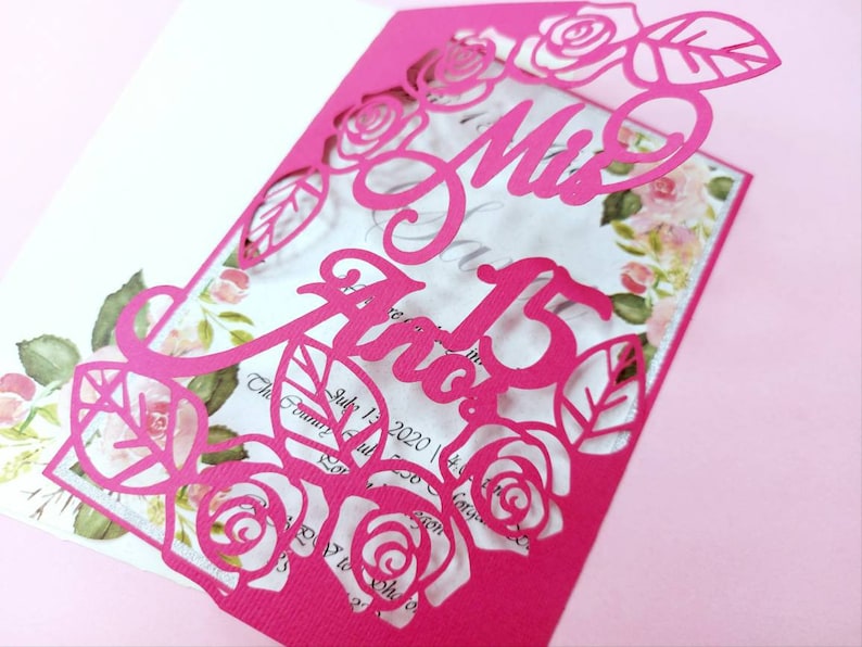 May include: A pink paper invitation with a cut-out design of roses and leaves. The text "Mis Quince A&ntilde;os" is written in a decorative font.
