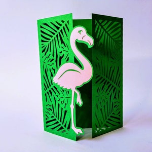 May include: A green card with a pink flamingo cut out in the center. The flamingo is standing on one leg and has its head turned to the side. The card is decorated with a leafy pattern.