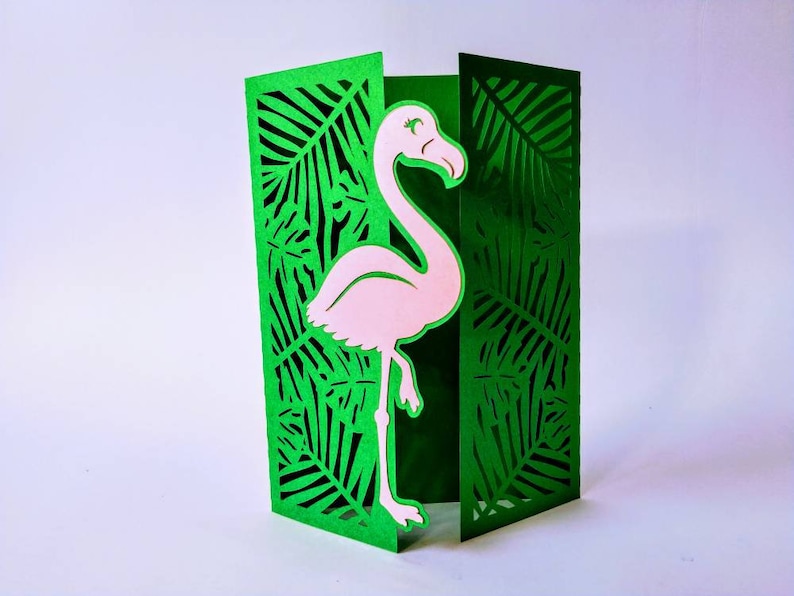 May include: A green card with a pink flamingo cut out in the center. The flamingo is standing on one leg and has its head turned to the side. The card is decorated with a leafy pattern.