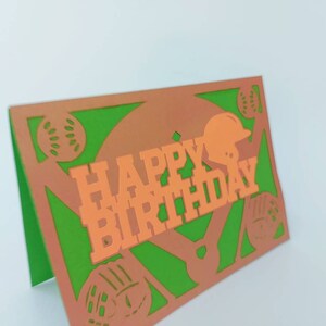 May include: A green and orange paper card with a baseball theme. The card says "Happy Birthday" in orange letters with a baseball helmet and baseballs cut out of the card.