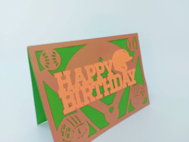 May include: A green and orange paper card with a baseball theme. The card says "Happy Birthday" in orange letters with a baseball helmet and baseballs cut out of the card.