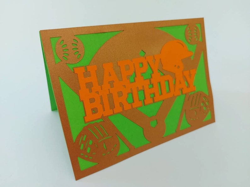 May include: A green and orange paper card with a baseball theme. The card says "Happy Birthday" in orange letters. There are baseballs and a baseball helmet cut out of the card.