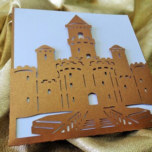 May include: A brown paper castle cutout with a white background. The castle has a tall central tower with two smaller towers on either side. The castle has a gate and a drawbridge.