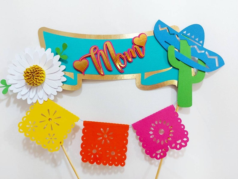 May include: A colorful paper craft set for a celebration. It includes a banner with the word "Mom" in gold letters, a cactus with a blue sombrero, a white daisy, and a string of colorful paper flags.
